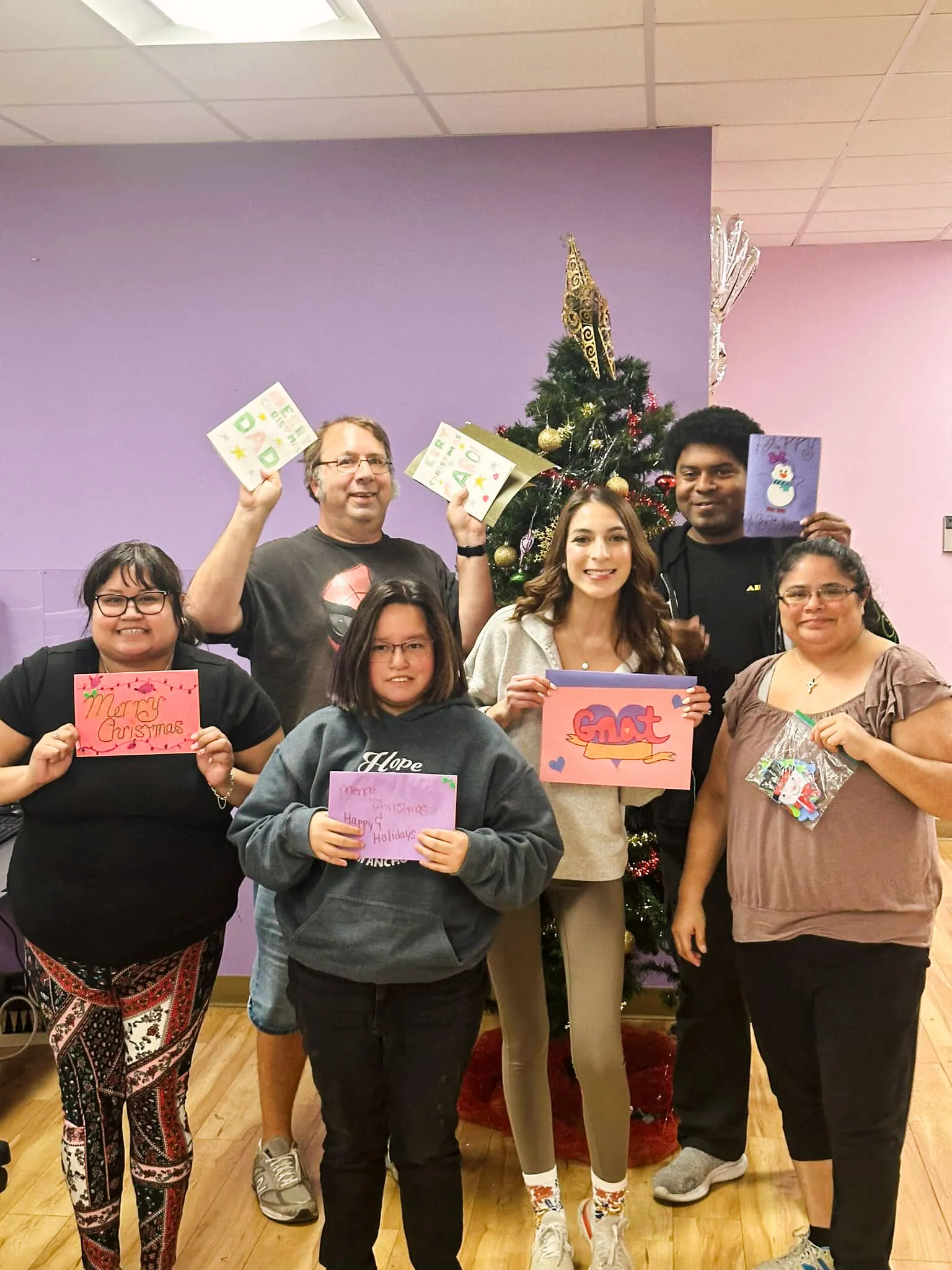Miembros y personal de San Antonio Clubhouse sonriendo con tarjetas navideñas hechas a mano frente a un árbol de Navidad